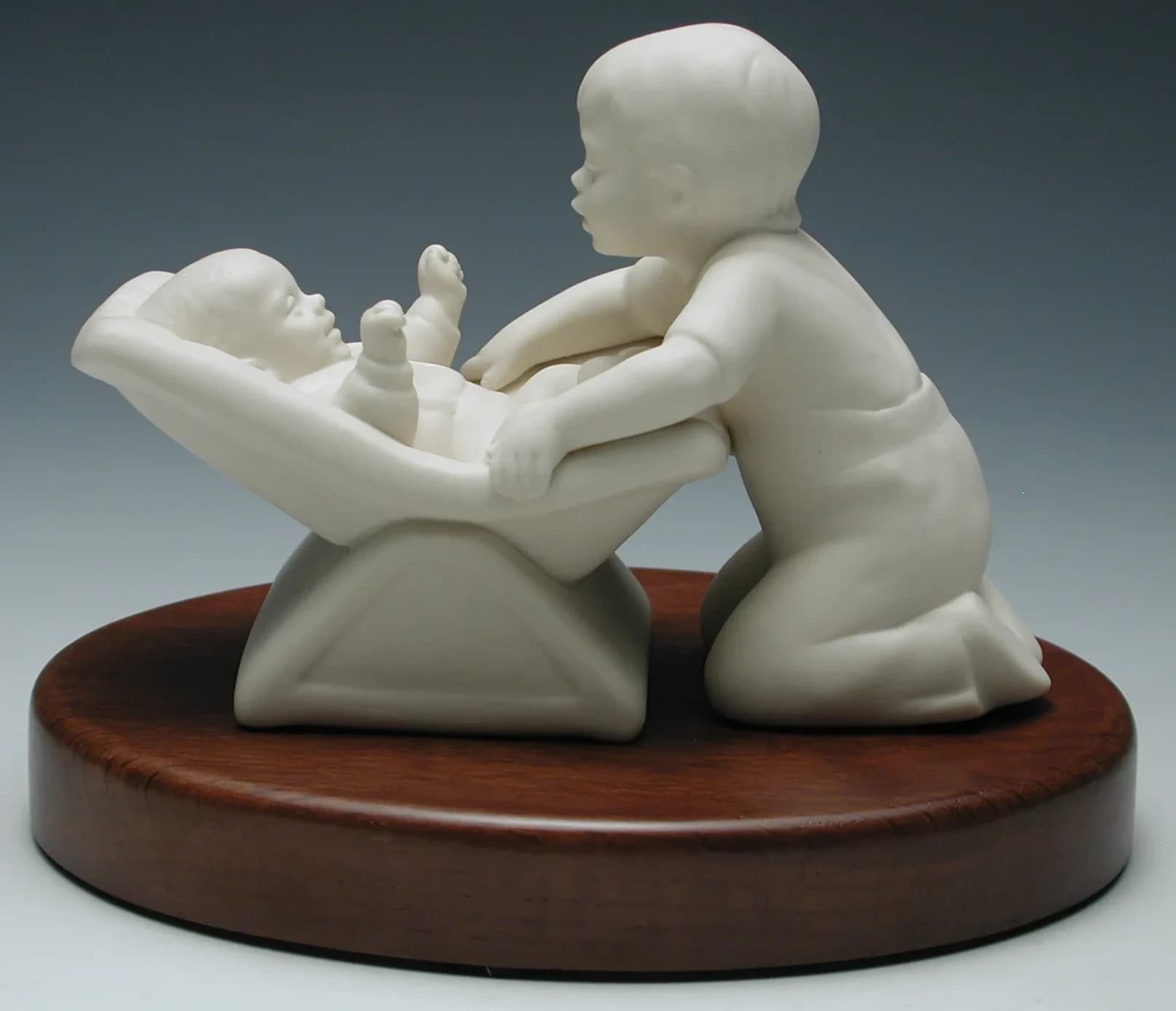 White porcelain figurine of a small boy kneeling to kiss a baby lying in a rocker, on an oval timber base