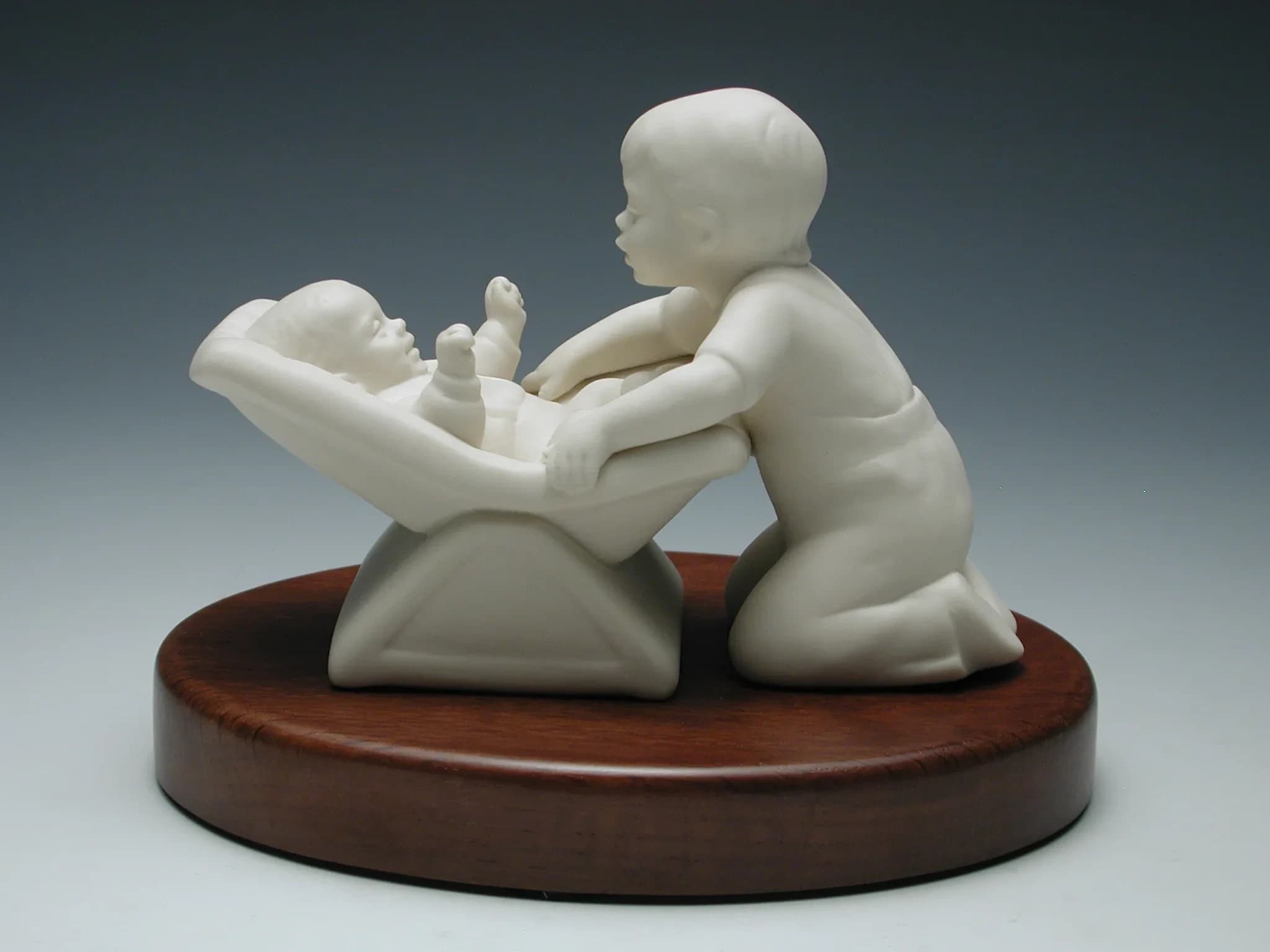 White porcelain figurine of a small boy kneeling to kiss a baby lying in a rocker, on an oval timber base