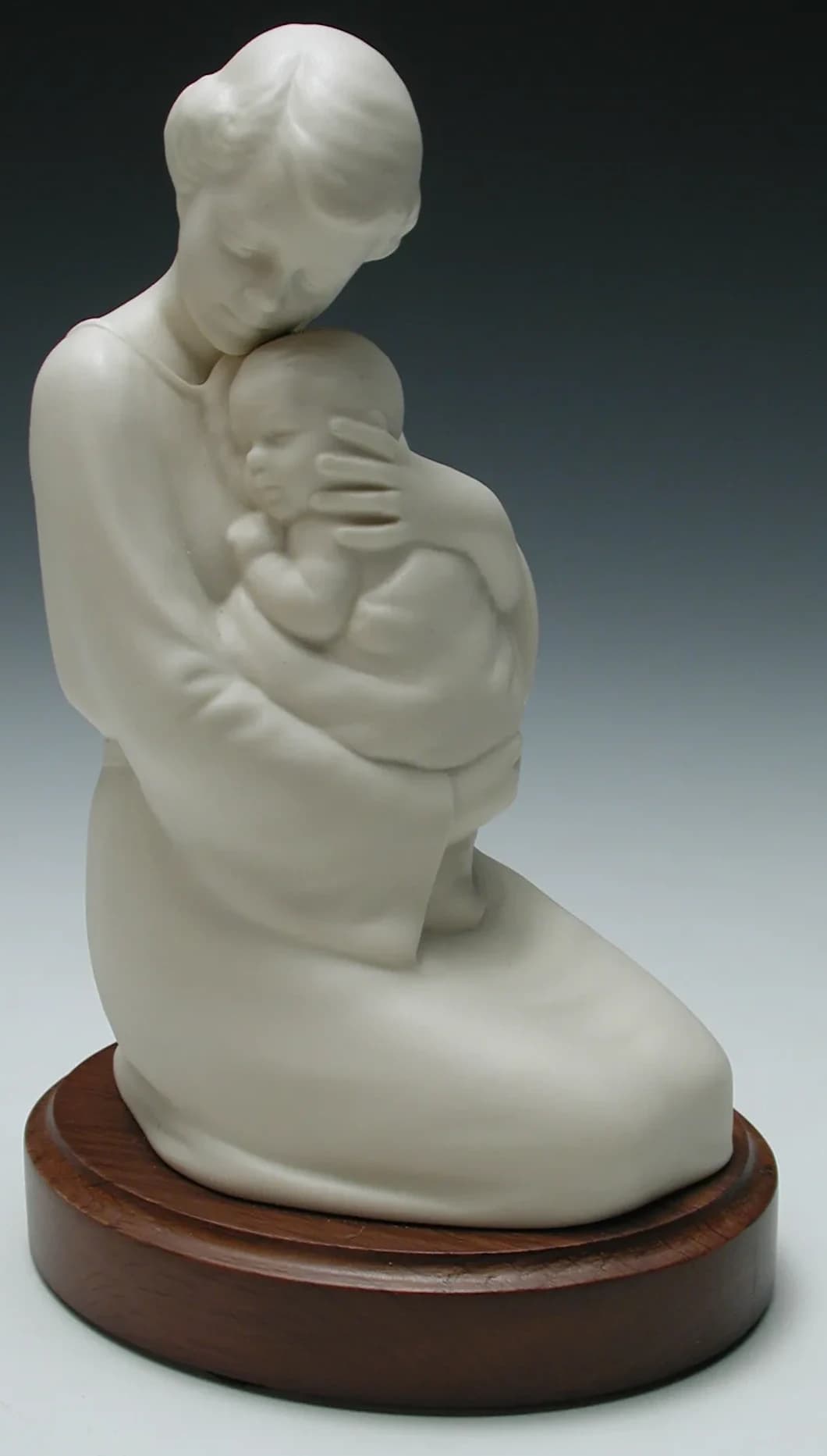 White porcelain figurine of a kneeling mother holding her sleeping baby close, on a round timber base