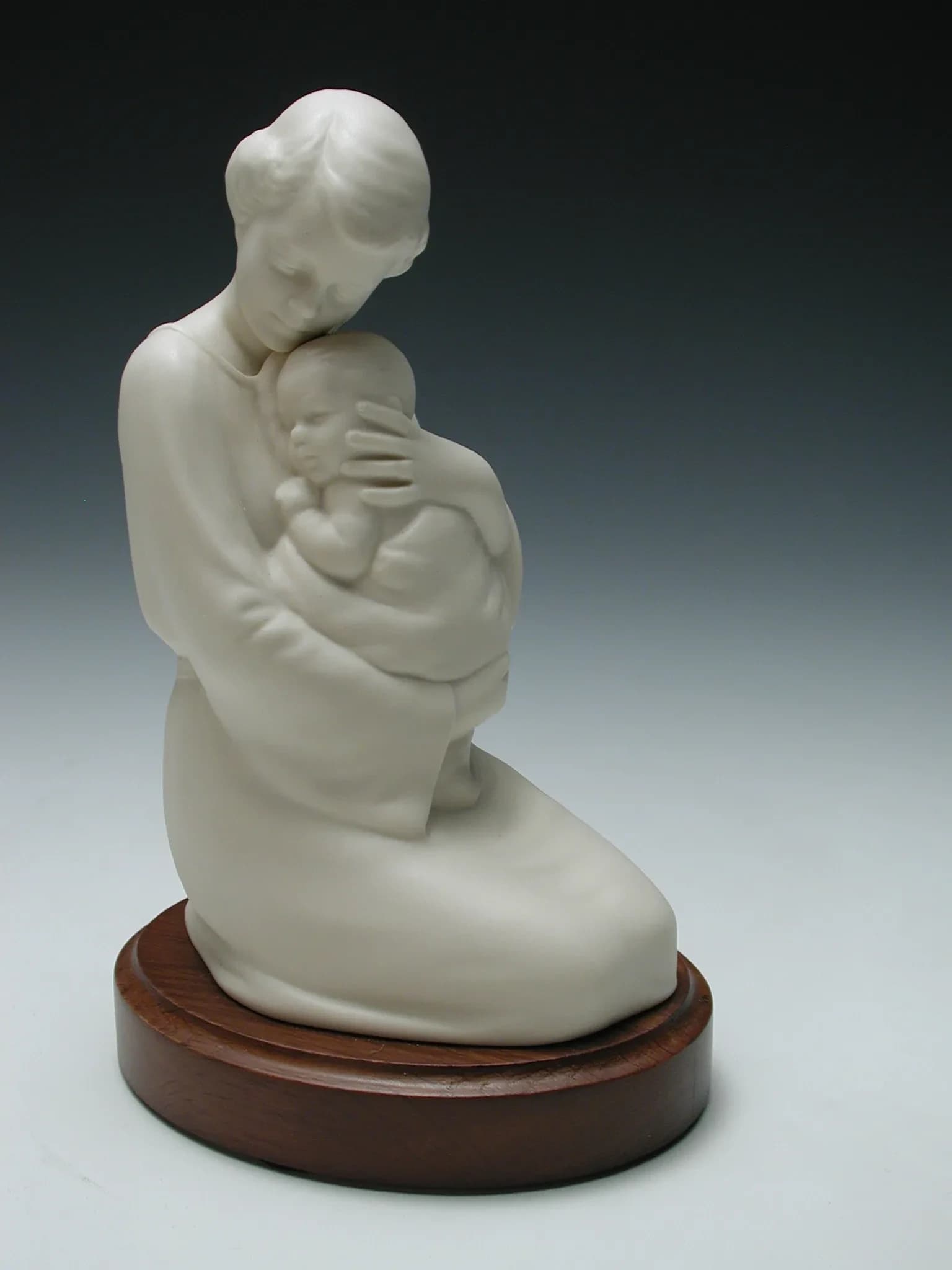 White porcelain figurine of a kneeling mother holding her sleeping baby close, on a round timber base