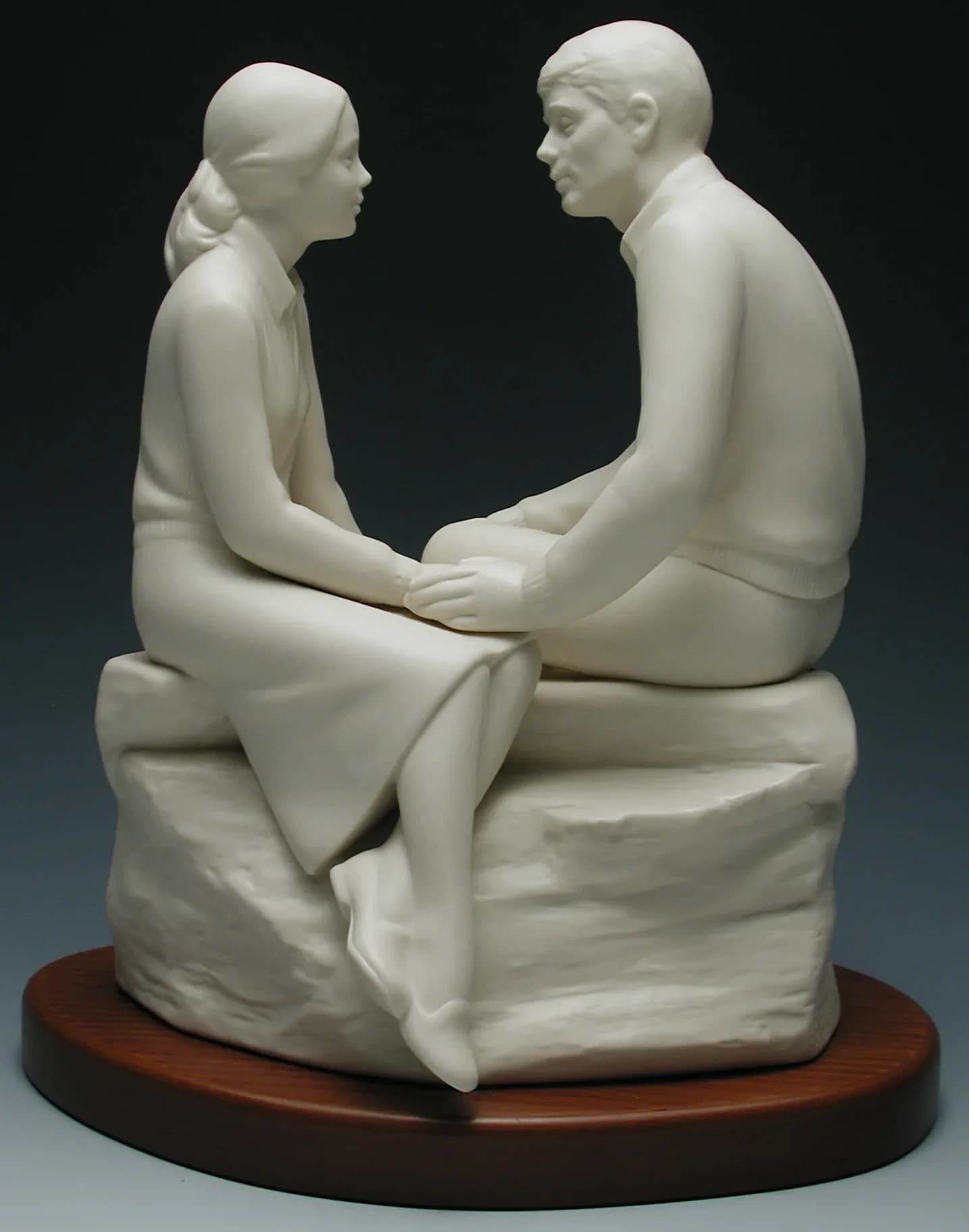 White porcelain figurine of a young couple sitting on a rock holding hands and gazing at each other, on an oval timber base