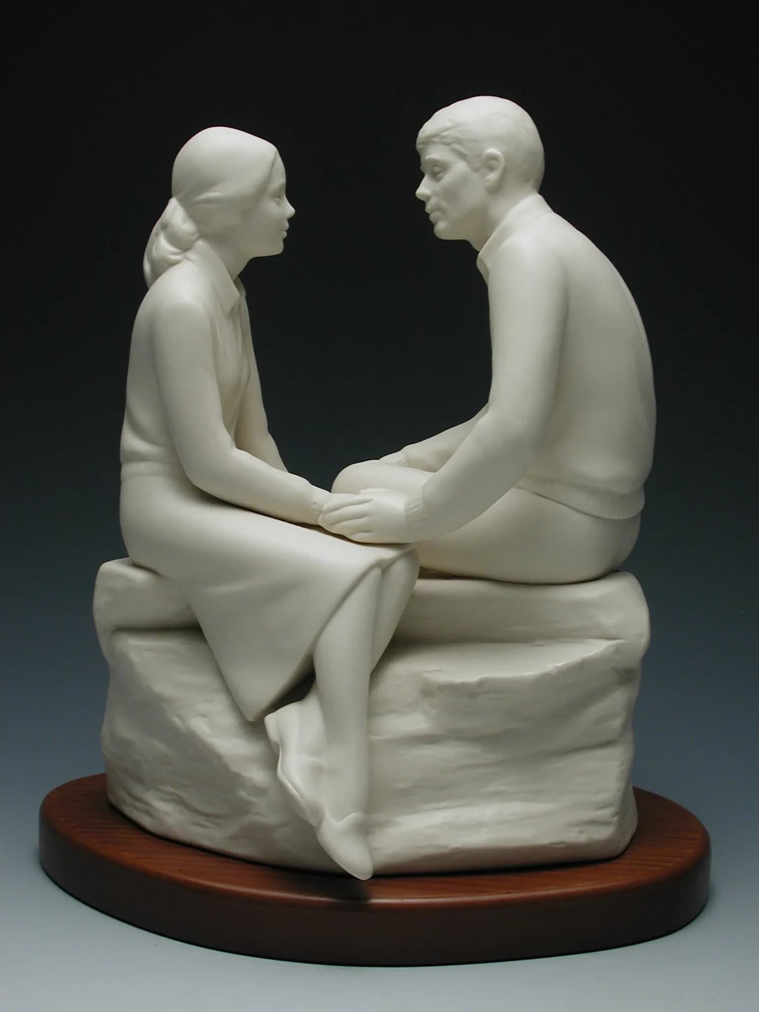 White porcelain figurine of a young couple sitting on a rock holding hands and gazing at each other, on an oval timber base