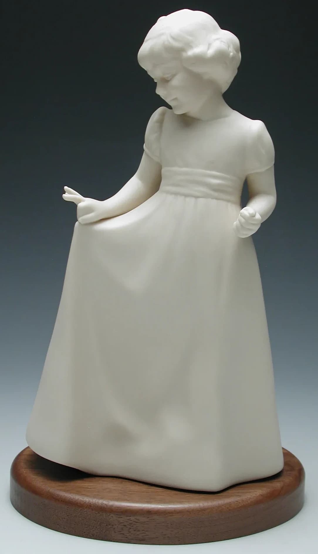 White porcelain figurine of a little girl in a long dress, holding out her skirt and looking down, on a round timber base