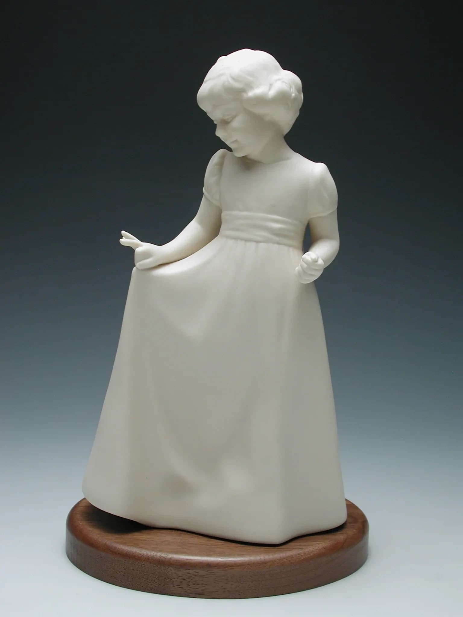White porcelain figurine of a little girl in a long dress, holding out her skirt and looking down, on a round timber base