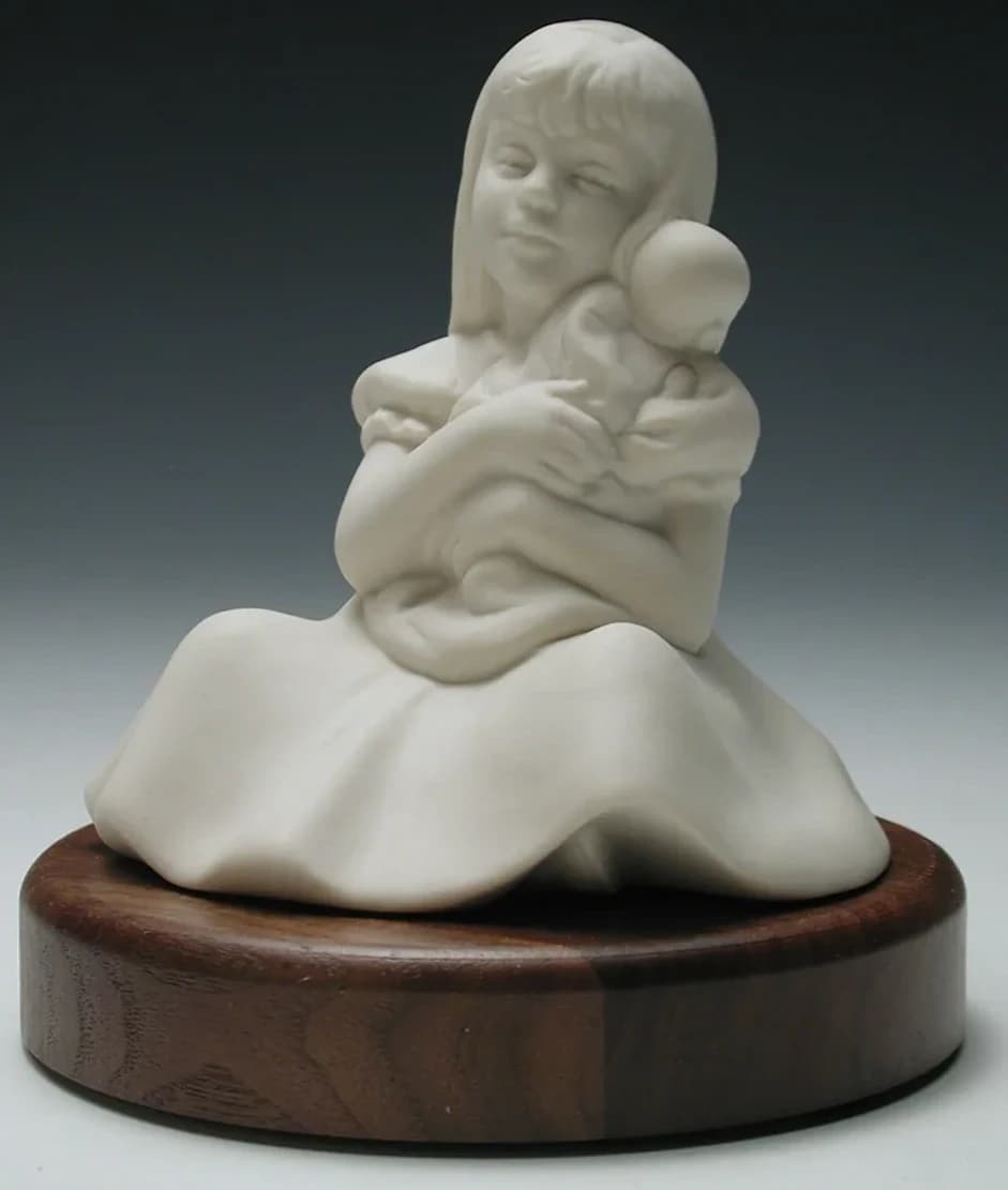 White porcelain figurine of a young girl sitting in a full skirt, hugging a doll to her cheek, on a round timber base